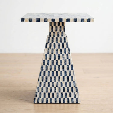 A accent side table with a rectangular top and tapered base, covered in a blue and owhite checkerboard capiz-inlay pattern, set on a light wood floor against a white wall.