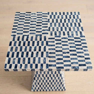 A square pedestal side table with a woven capiz-style pattern in dark blue and off-white, set against a light wood floor background.