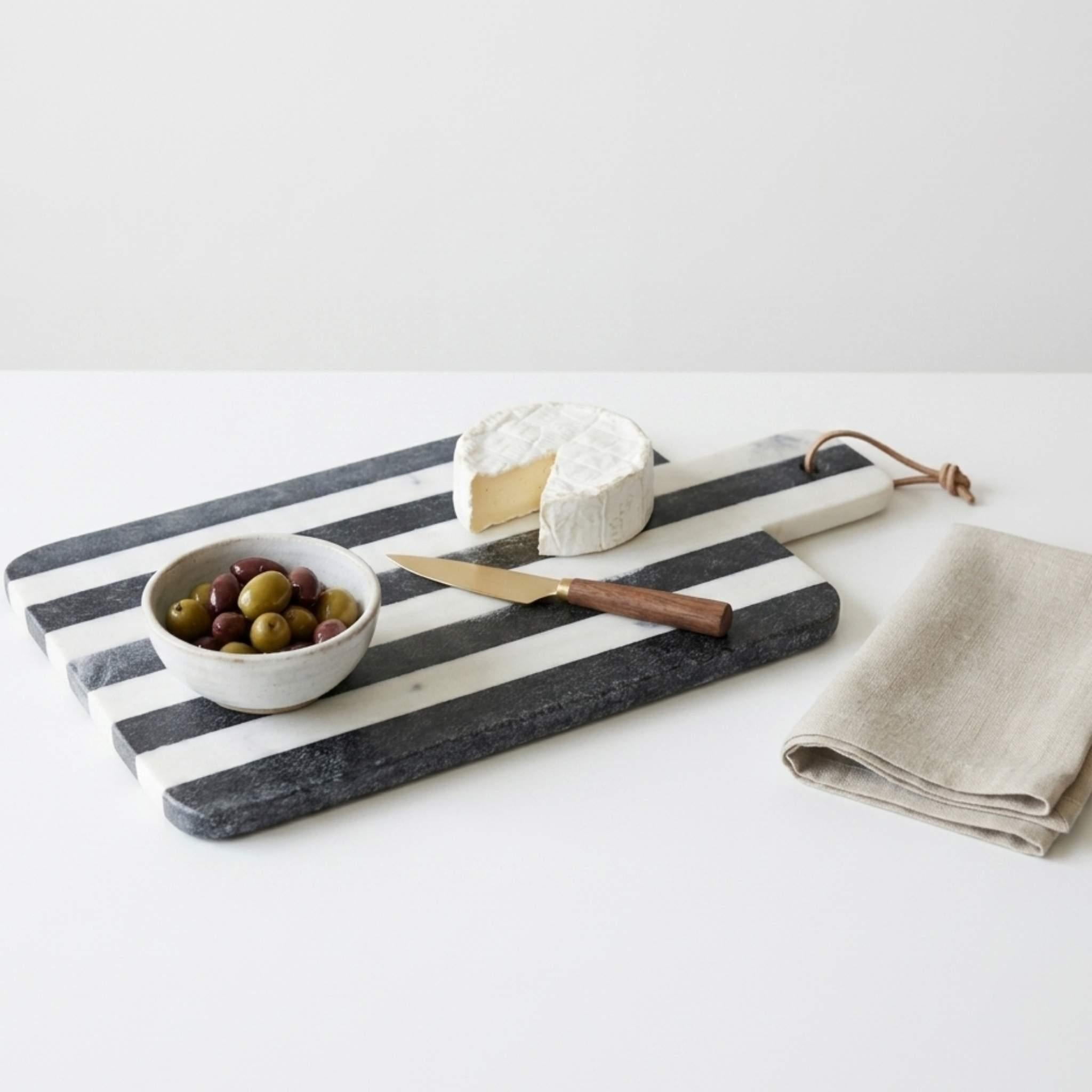 Black and white striped marble cutting board with a bowl of olives, cheese, and a knife on a white surface.