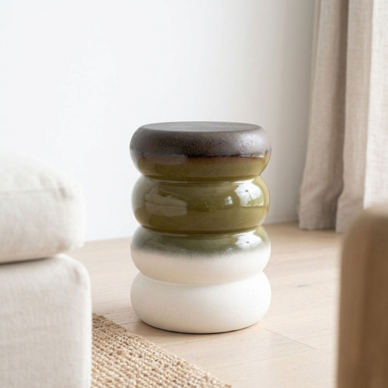A small round ceramic stool with a stacked, ribbed design in earthy tones of olive green, cream, and dark brown, set in living room.