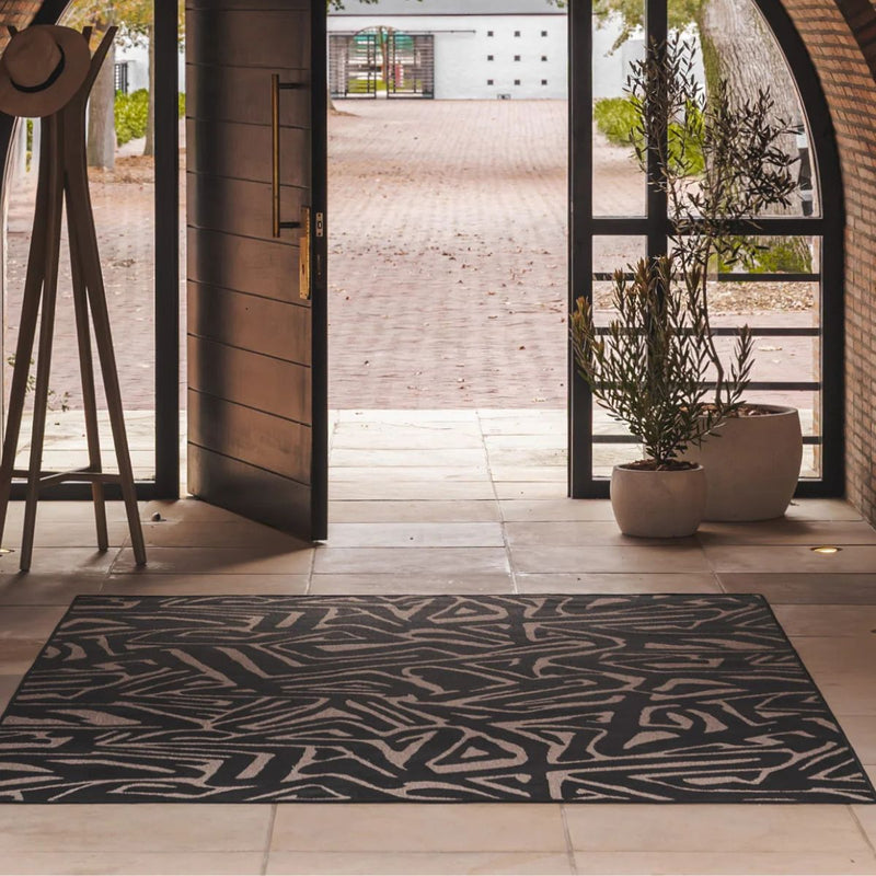 Area rug with an abstract black and beige geometric pattern, on a tiled entryway floor in front of open glass doors.