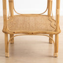 Close-up of a light natural rattan and wicker side table with woven cane backing and curved wicker trim, shown on a pale wood floor against a white background.