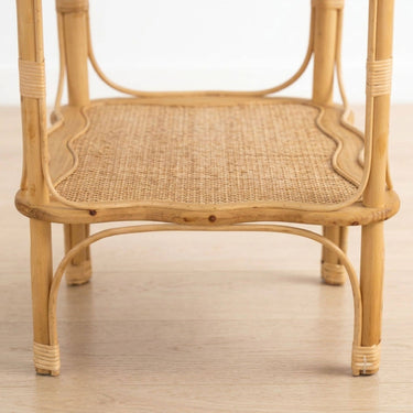 Close-up of a light natural rattan and wicker side table with woven cane backing and curved wicker trim, shown on a pale wood floor against a white background.