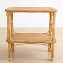 A two-tier rattan side table with a natural light brown finish, featuring woven cane panels on the top and middle shelves, set against a plain white wall and light wood floor.
