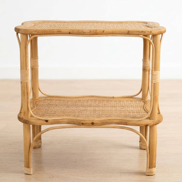 A two-tier rattan side table with a natural light brown finish, featuring woven cane panels on the top and middle shelves, set against a plain white wall and light wood floor.